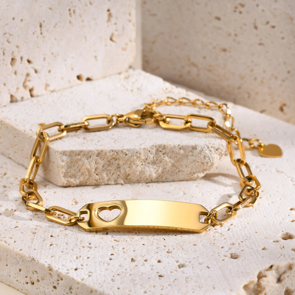 A gold-plated ID bracelet with a rectangular bar featuring a heart-shaped cutout, displayed on a textured beige stone block.