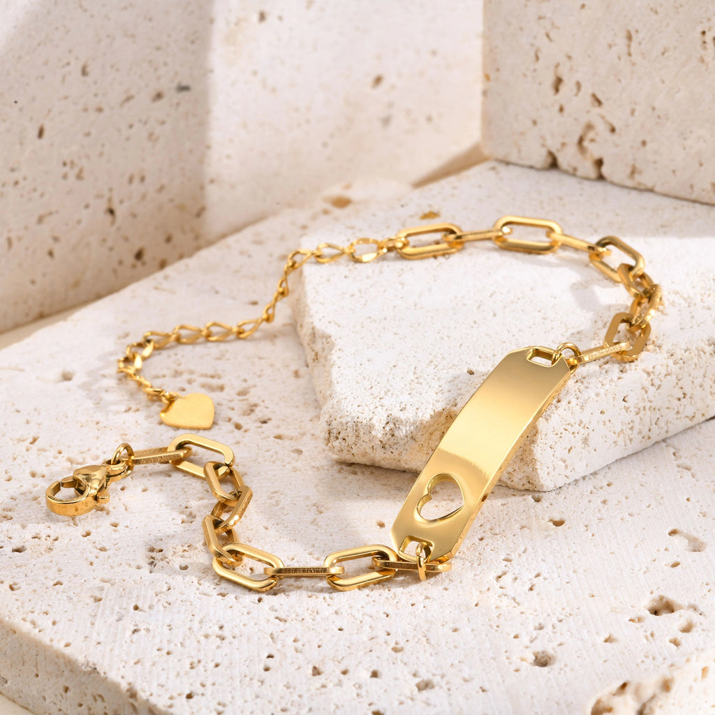 A gold-plated ID bracelet with a rectangular bar, heart-shaped cutout, and link chain, displayed on textured beige stone blocks.