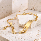 A gold-plated ID bracelet with a rectangular bar, heart-shaped cutout, and link chain, displayed on textured beige stone blocks.