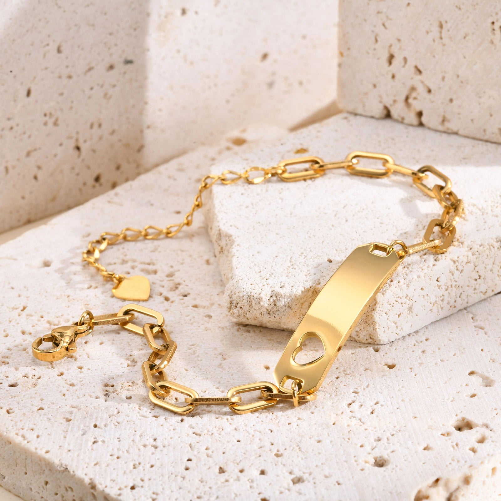 A gold-plated ID bracelet with a rectangular bar, heart-shaped cutout, and link chain, displayed on textured beige stone blocks.