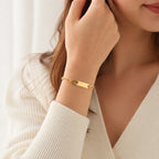 A close-up lifestyle shot of a woman wearing a gold-plated ID bracelet on her wrist, paired with a cream-colored ribbed knit cardigan.