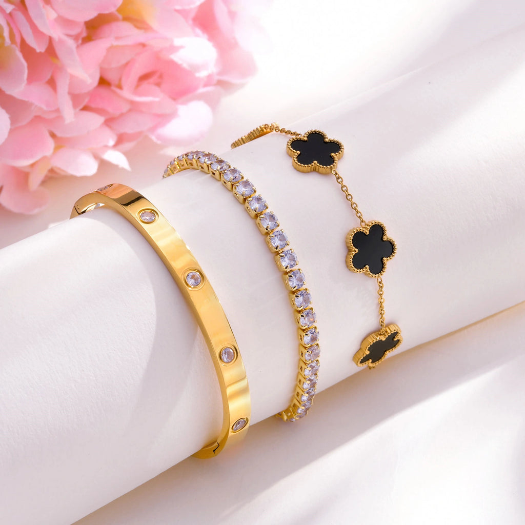 Black and gold floral bracelet set displayed on a soft white surface with pink flowers, featuring a black flower chain bracelet, CZ tennis bracelet, and gold tone bangle.