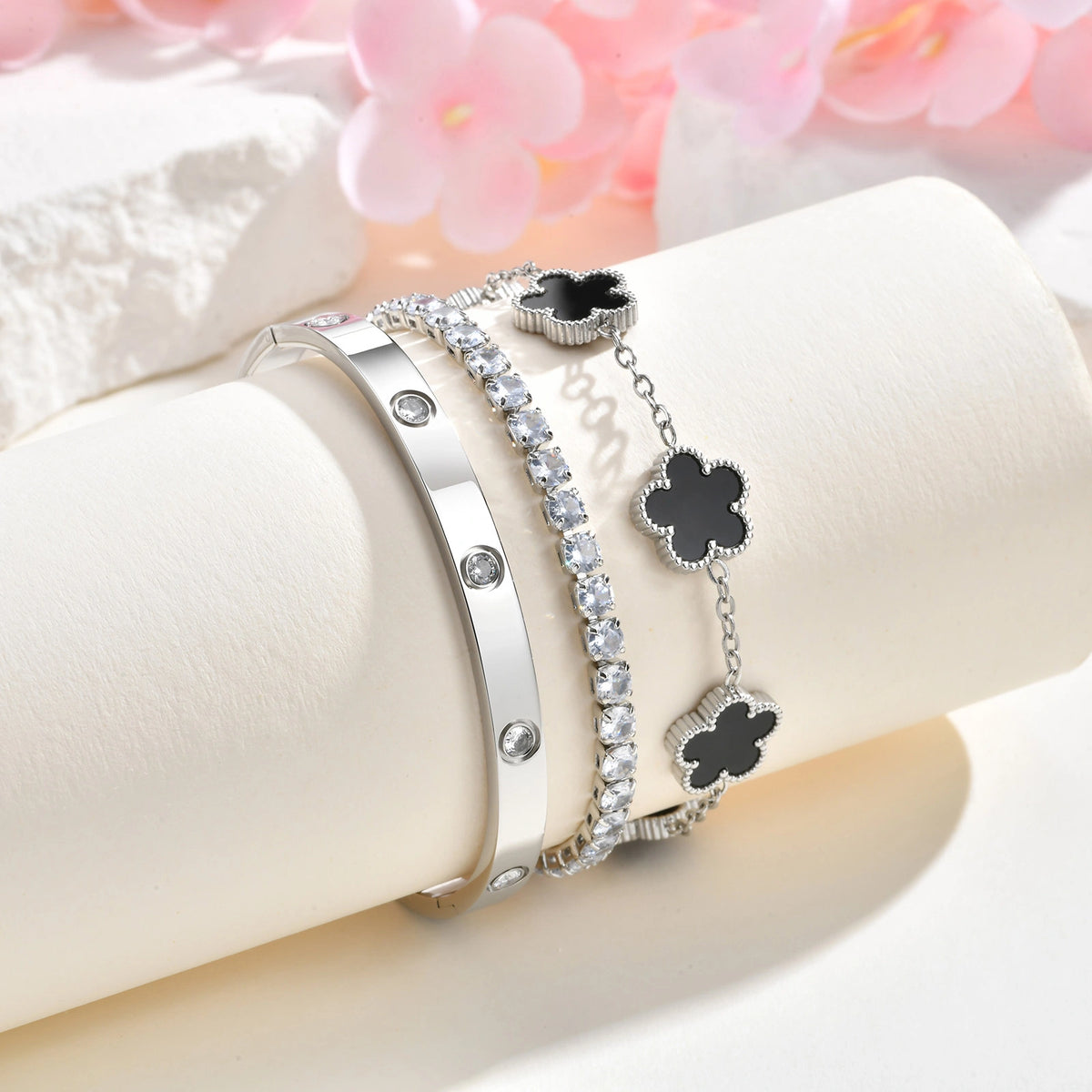 Silver tone bracelet stack displayed on a white jewelry stand with pink flowers, featuring black mother of pearl charms, a CZ tennis bracelet, and a crystal bangle.