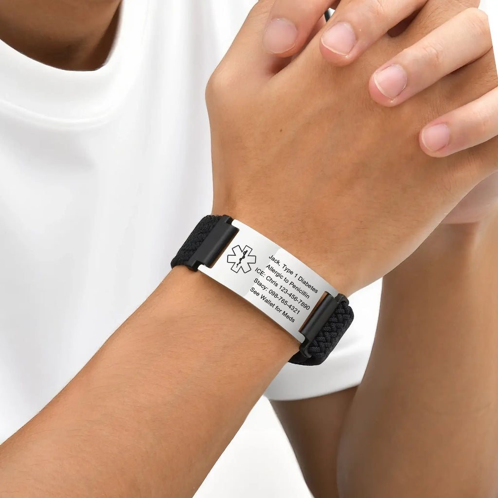 Person wearing a black braided medical ID bracelet with a curved stainless steel ID plate on the wrist.