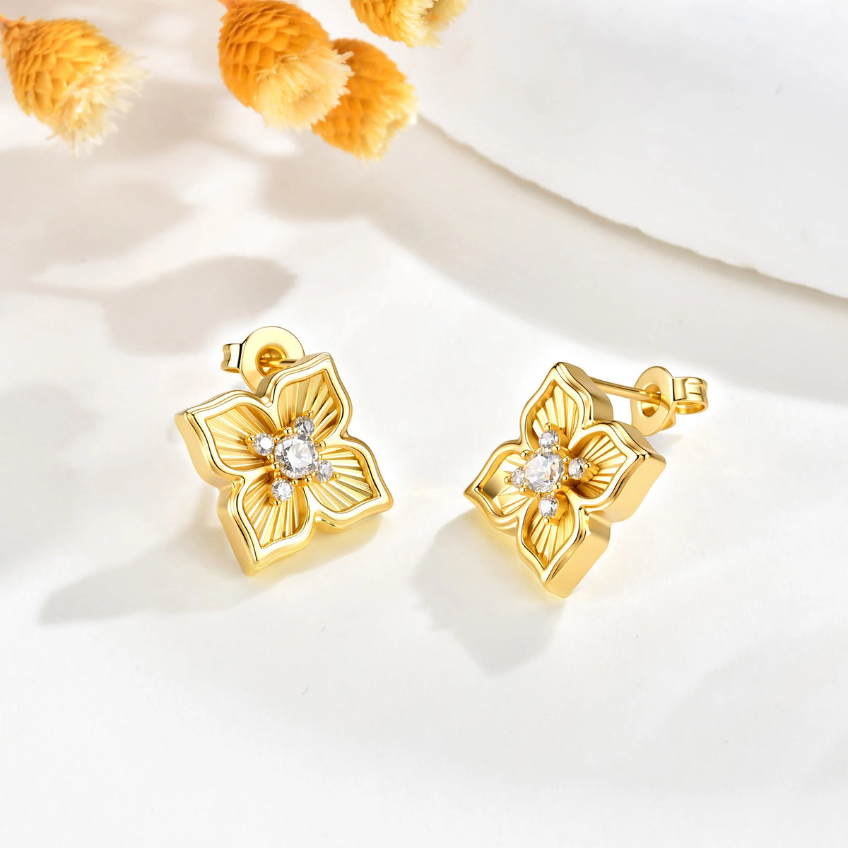 Pair of gold four leaf clover CZ stud earrings displayed on a white surface with delicate yellow floral accents.