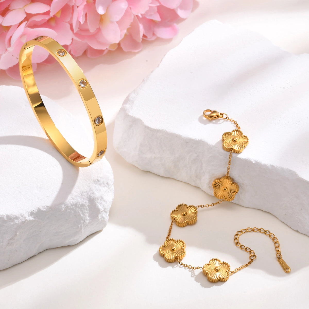 Gold tone diamond-cut flower bracelet and crystal bangle displayed on white jewelry props with soft pink flowers in the background.