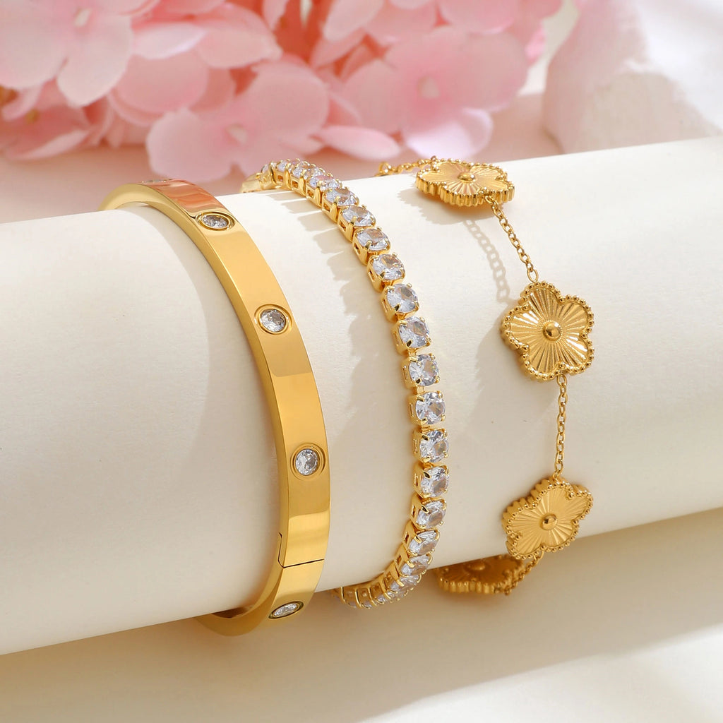 Gold tone floral bracelet stack displayed on a white jewelry stand beside pink flowers, featuring gold flower charms, a CZ tennis bracelet, and a crystal bangle.