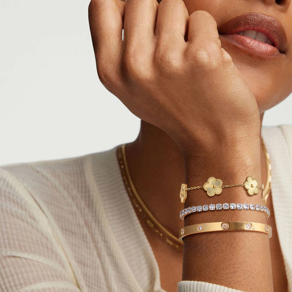 Close-up of a model wearing layered gold tone bracelets with textured gold flower charms, a sparkling CZ tennis bracelet, and a polished bangle.