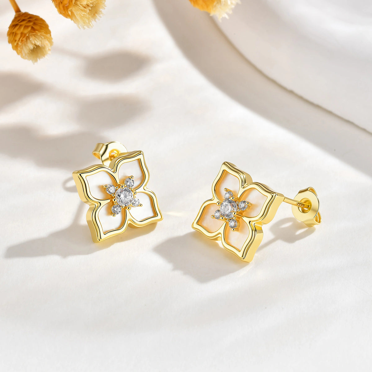 Pair of gold mother of pearl floral stud earrings displayed on a soft white surface with delicate yellow flowers.