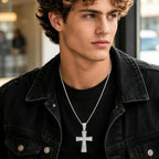 Male model wearing a silver tone iced out cross pendant necklace with a rope chain, styled with a black jacket for a modern streetwear look.