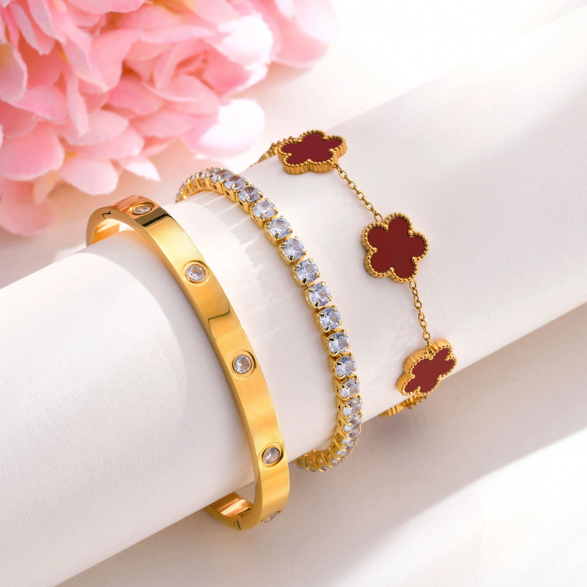 Red and gold floral bracelet set displayed on a white jewelry stand beside pink flowers, featuring a red flower bracelet, CZ tennis bracelet, and crystal bangle.