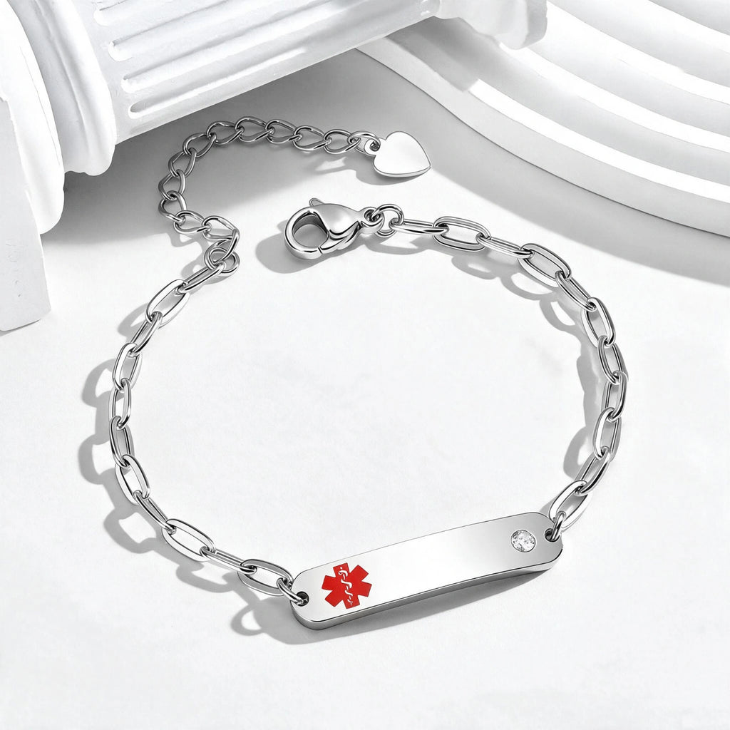 A silver-tone ID bracelet featuring a flat bar engraved with a red medical star of life symbol and a small clear rhinestone, displayed on a clean white surface with decorative white columns.