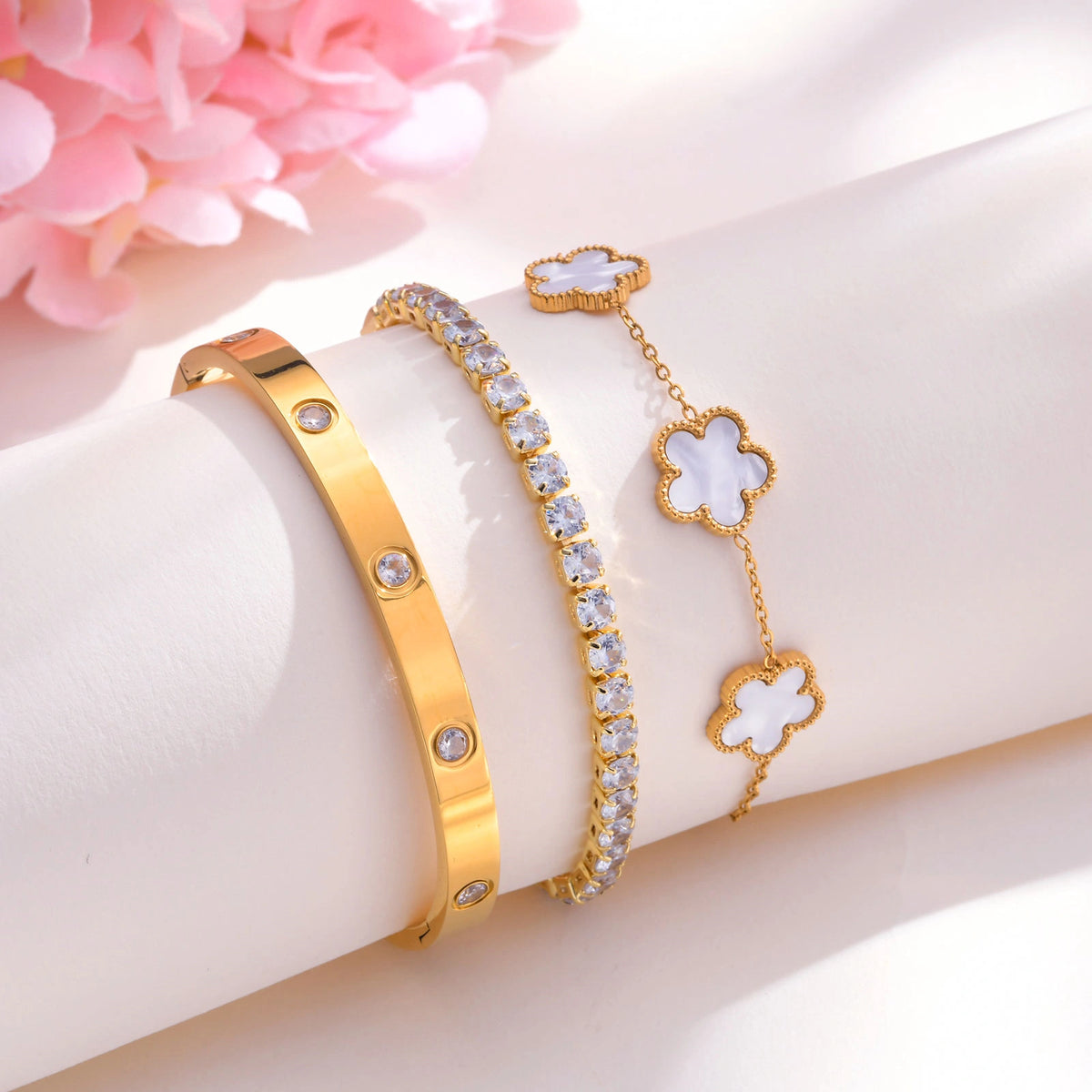 Gold tone bracelet stack featuring a white mother of pearl floral chain bracelet, cubic zirconia tennis bracelet, and crystal bangle displayed beside pink flowers.
