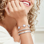 Close-up of a woman wearing layered silver tone bracelets with white mother of pearl flower charms, sparkling cubic zirconia stones, and a polished crystal bangle.