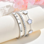 Silver tone bracelet stack displayed on a white jewelry stand beside pink flowers, featuring white mother of pearl floral charms, a CZ tennis bracelet, and a crystal bangle.