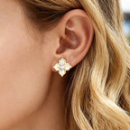 Close-up of a woman wearing gold four leaf clover stud earrings with white mother of pearl petals and sparkling cubic zirconia accents.