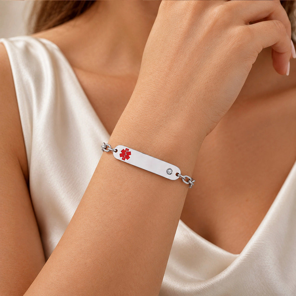 A silver-toned metal ID bracelet with a red medical cross symbol, worn on a woman's wrist. The bracelet is shown on a model wearing a white satin top.