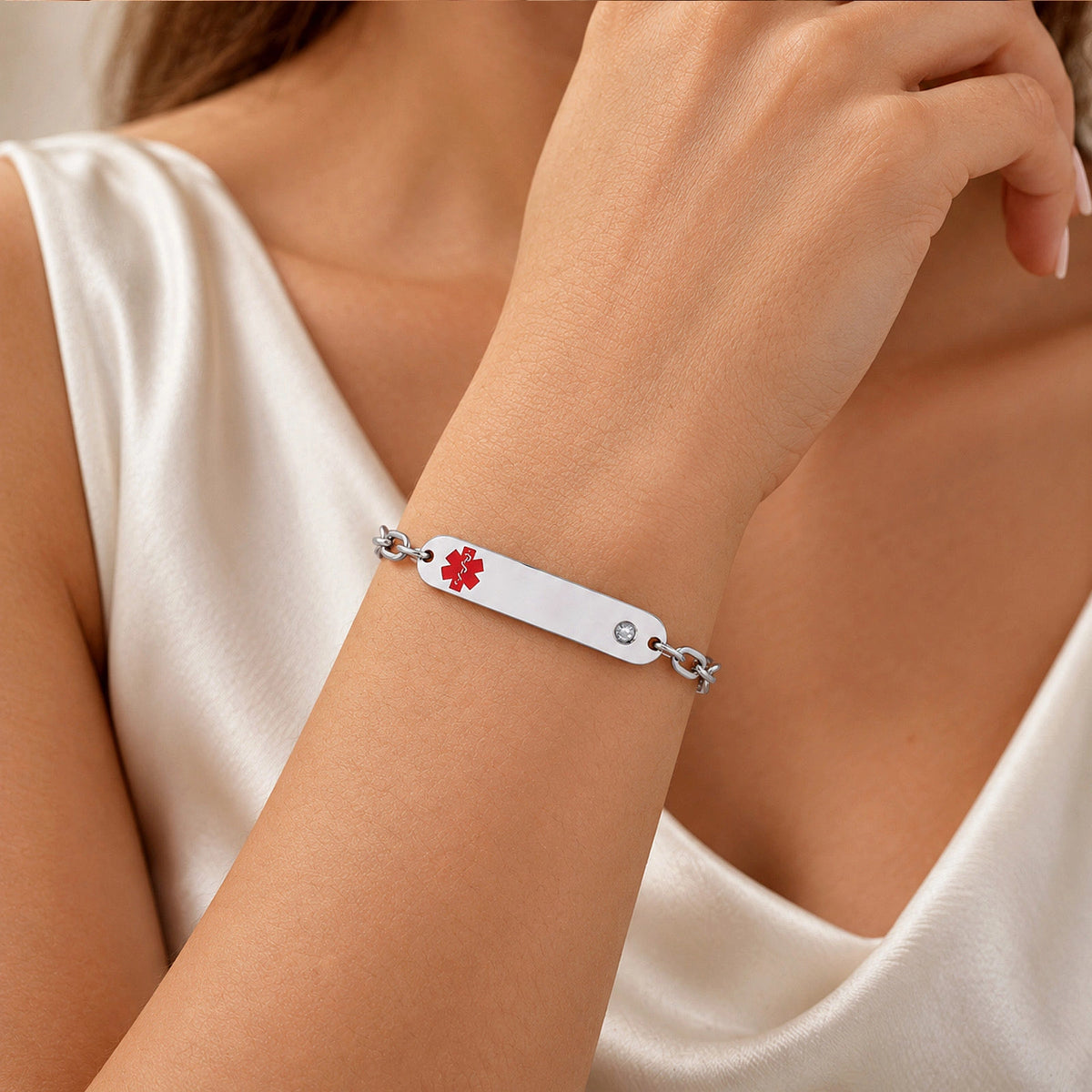 A silver-toned metal ID bracelet with a red medical cross symbol, worn on a woman's wrist. The bracelet is shown on a model wearing a white satin top.